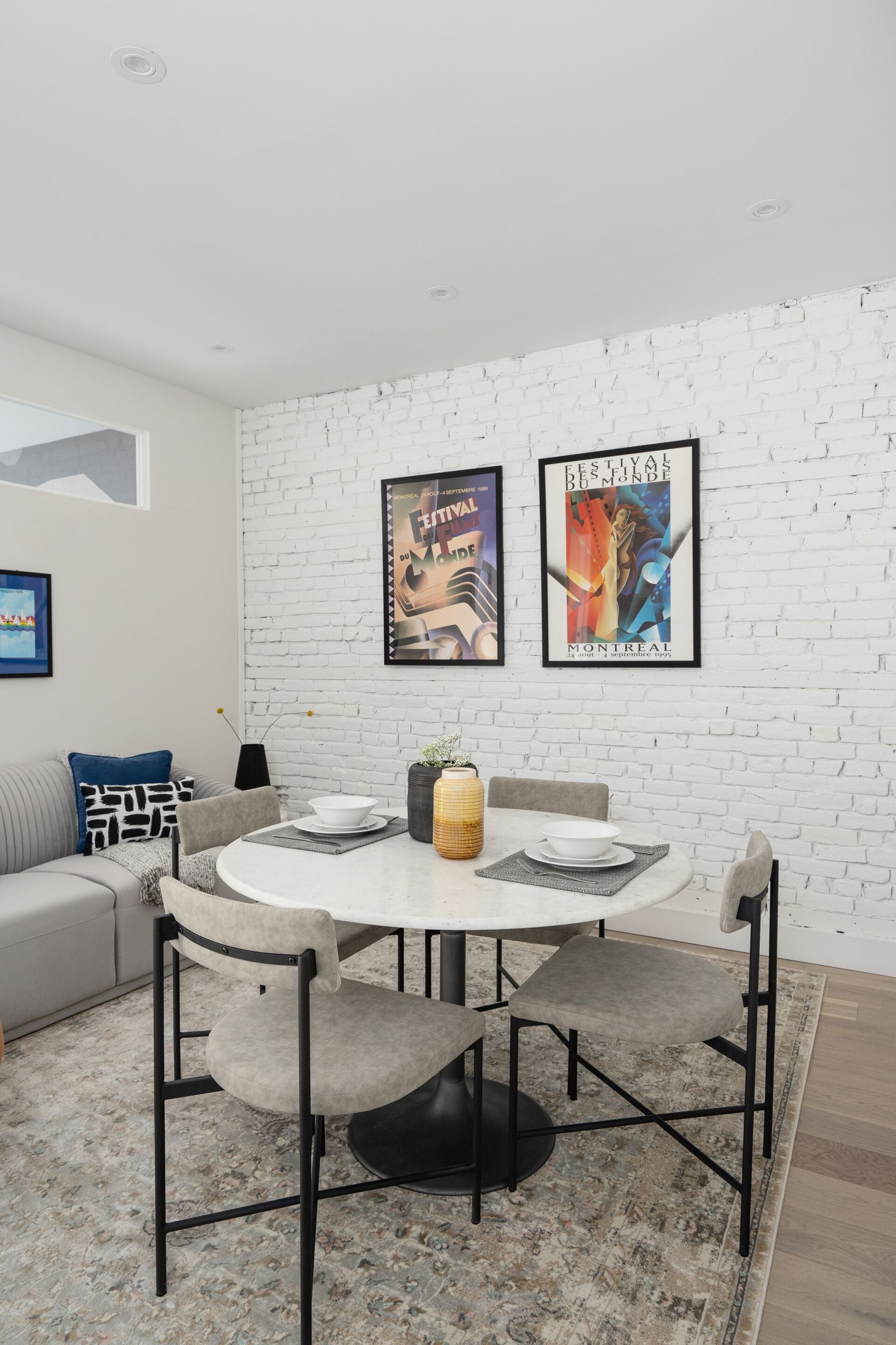 Un comedor moderno con una mesa redonda, cuatro sillas y una pared de ladrillo blanco. Hay dos carteles enmarcados en la pared, un sofá gris y una alfombra estampada.