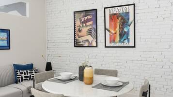 Un comedor moderno con una mesa redonda, cuatro sillas y una pared de ladrillo blanco. Hay dos carteles enmarcados en la pared, un sofá gris y una alfombra estampada.