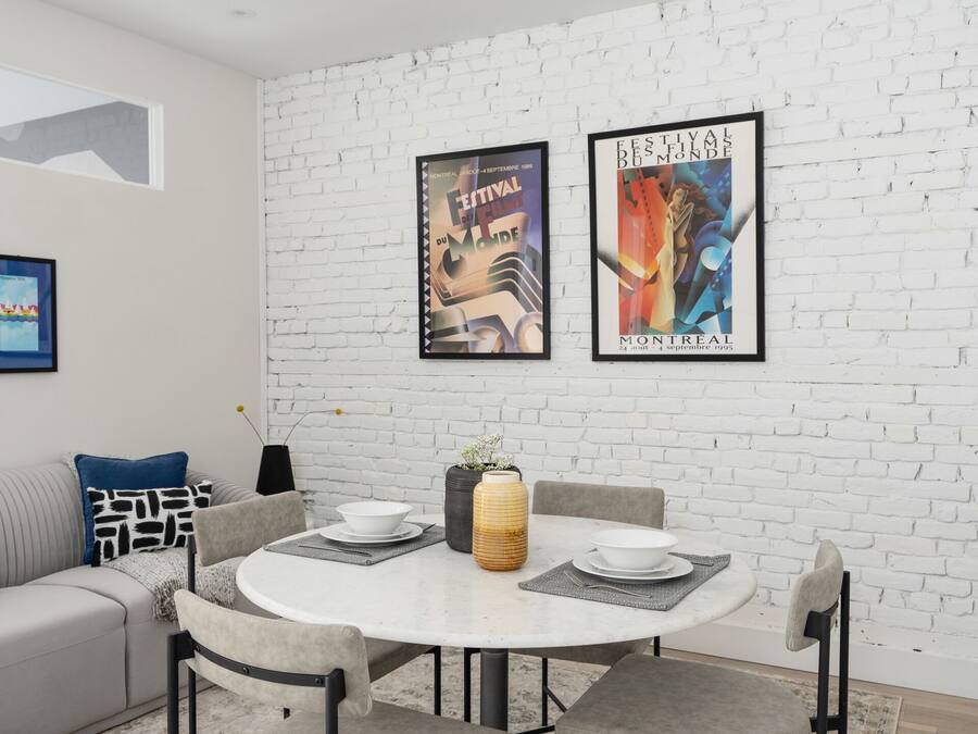 Un comedor moderno con una mesa redonda, cuatro sillas y una pared de ladrillo blanco. Hay dos carteles enmarcados en la pared, un sofá gris y una alfombra estampada.