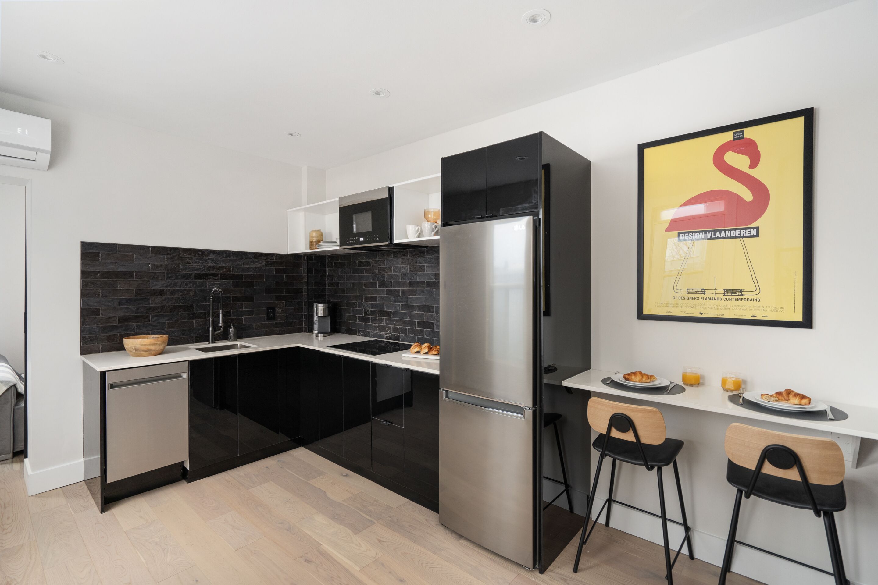 A modern kitchen with black cabinetry, a stainless steel refrigerator, and a black backsplash. There are two bar stools and a small dining table with plates of food and drinks.