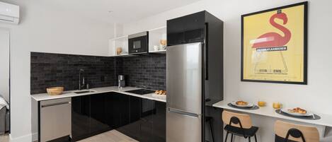 Una cocina moderna con armarios negros, un refrigerador de acero inoxidable y una cubierta trasera negra. Hay dos taburetes de bar y una pequeña mesa de comedor con platos de comida y bebidas.