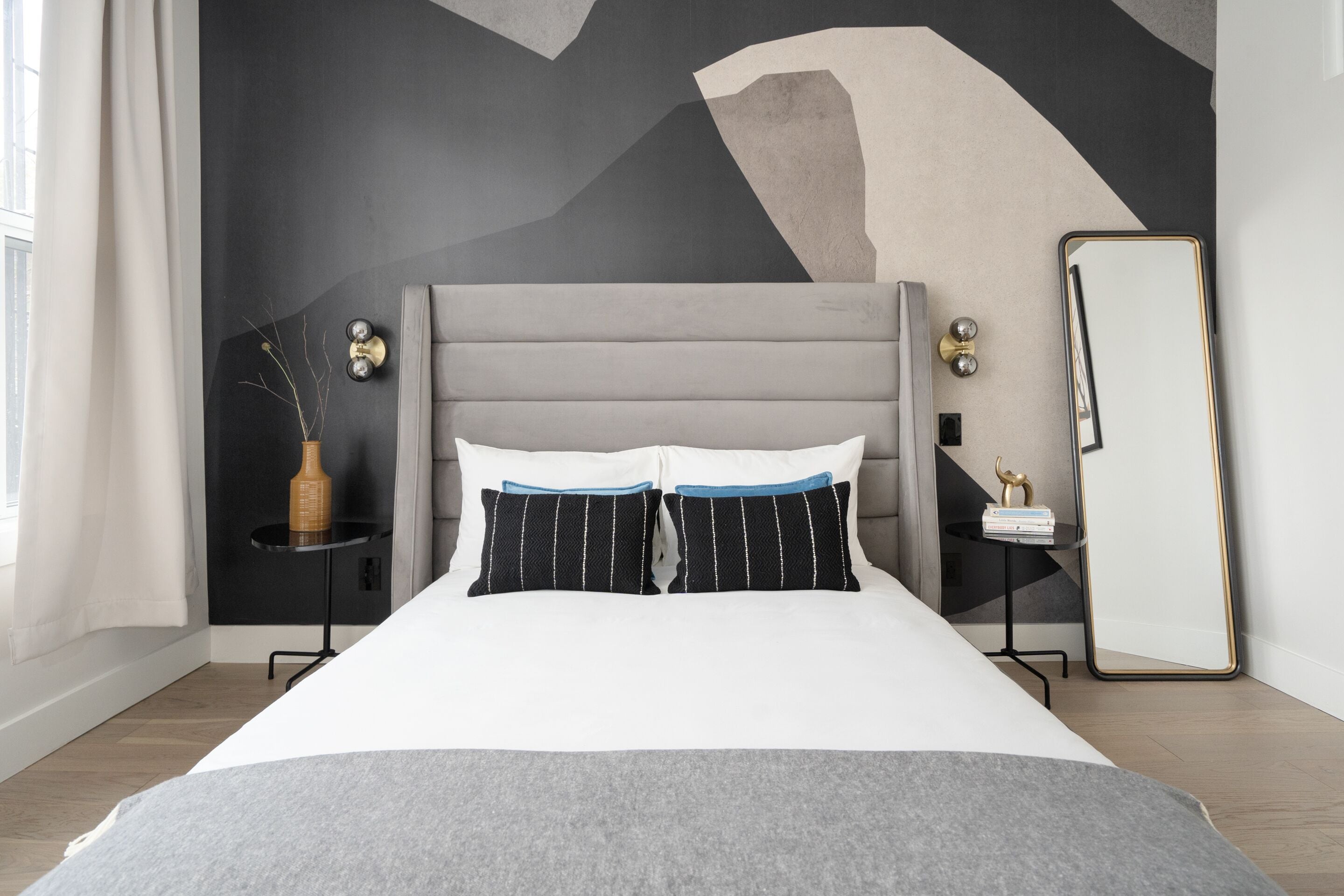 Een moderne slaapkamer met een grijs hoofdbord, wit beddengoed, een klein nachtkastje met een vaas en een grote spiegel.