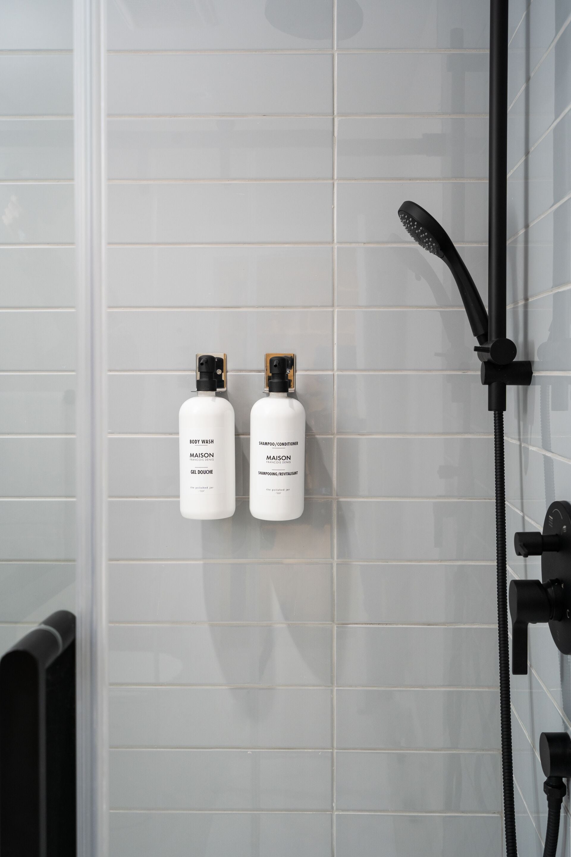 A bathroom with white tiled walls, a black showerhead, and two bottles of Maison body wash and shampoo.