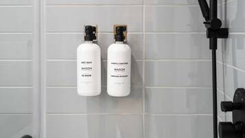 A bathroom with white tiled walls, a black showerhead, and two bottles of Maison body wash and shampoo.