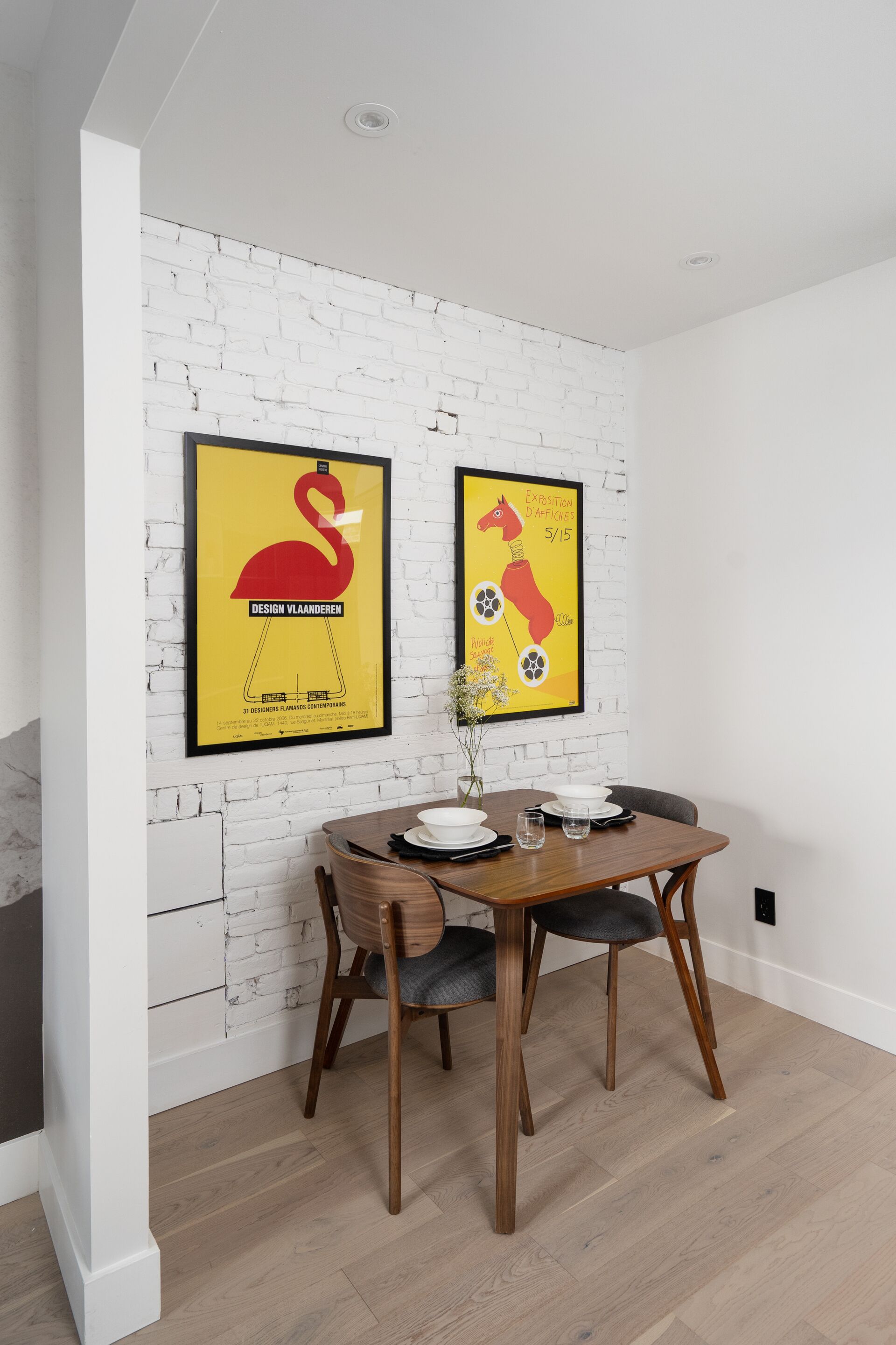A dining area with a white brick wall, two framed posters, a wooden dining table with four chairs, and a vase with flowers.
