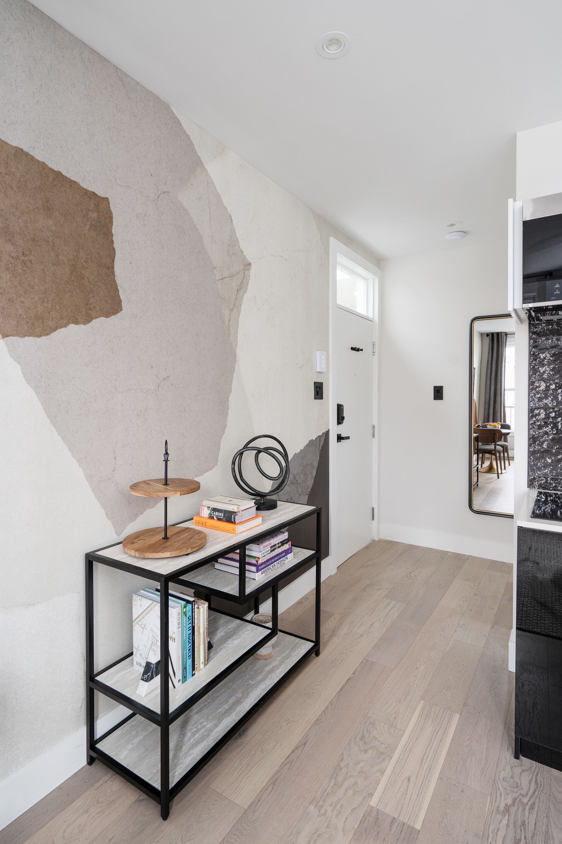 Un pasillo moderno con suelo de madera, una consola metálica negra con libros y un espejo, y una puerta blanca.