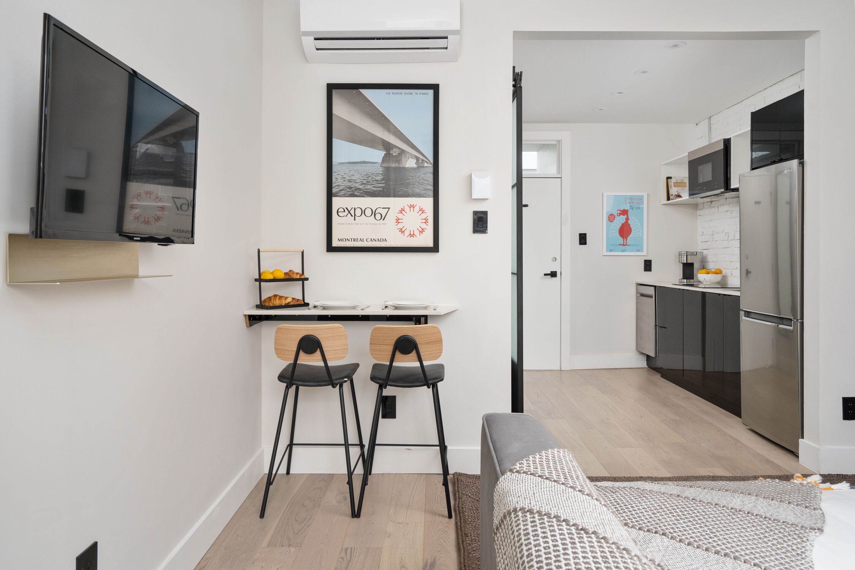 Una cocina moderna con refrigerador, microondas y estufa. Hay una zona de comedor con dos sillas y una mesa pequeña. Un televisor está montado en la pared, y se exhibe un cartel sobre la mesa.
