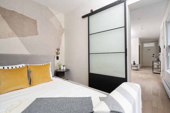 A modern bedroom with a bed, a sliding door, and a view into another room. - François-Denis Apartments (Montreal)