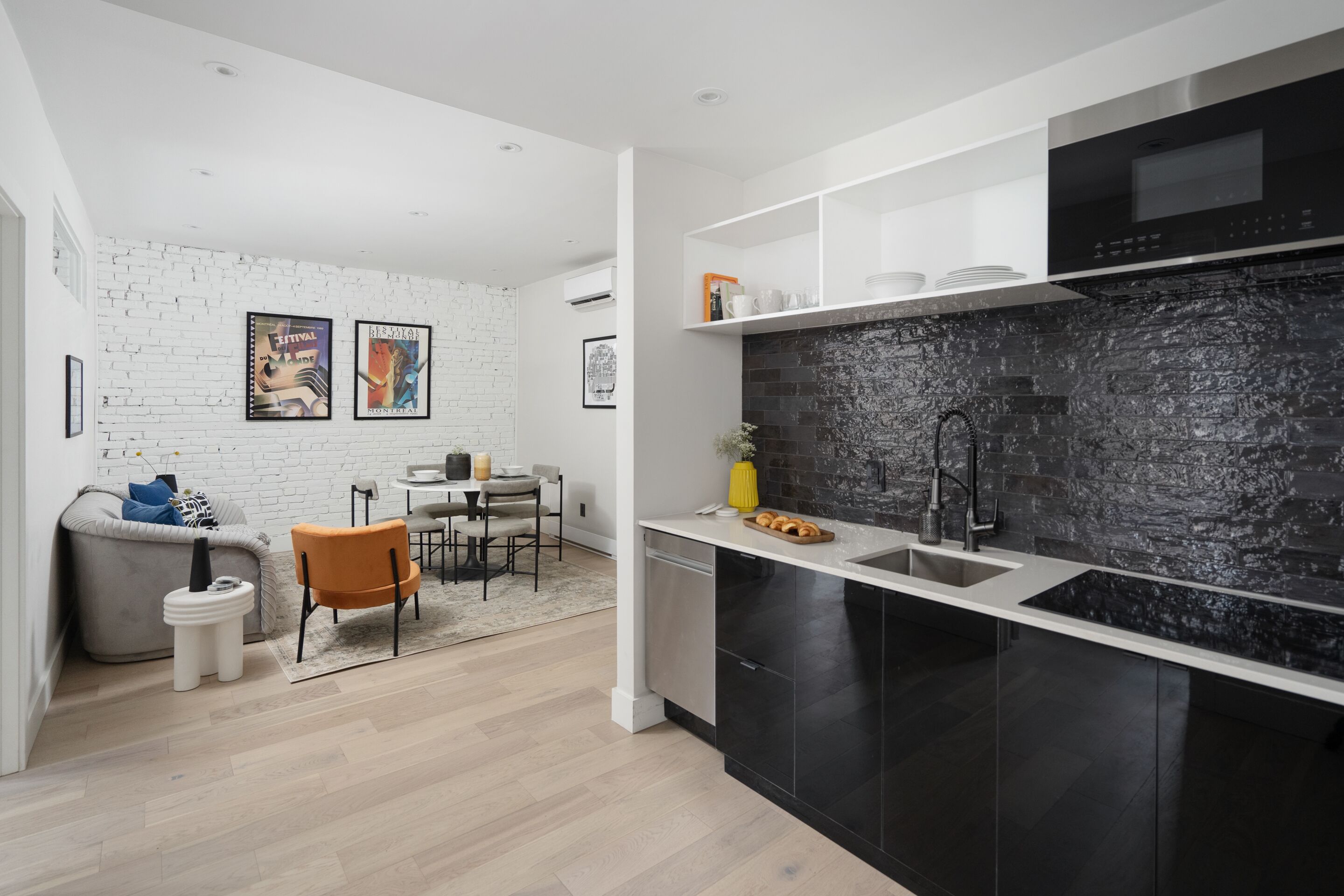 A modern kitchen with black cabinets, a white countertop, and a black backsplash. There is a dining area with a gray sofa and orange chairs.