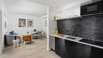 A modern kitchen with black cabinets, a white countertop, and a black backsplash. There is a dining area with a gray sofa and orange chairs.
