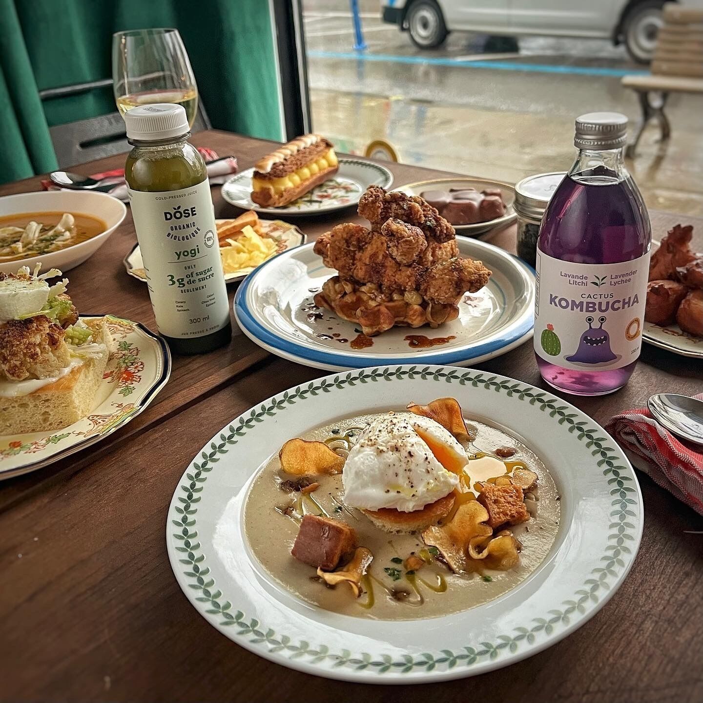 A table with various dishes, including a plate with a poached egg and mushrooms, a bottle of kombucha, and a bottle of organic juice.