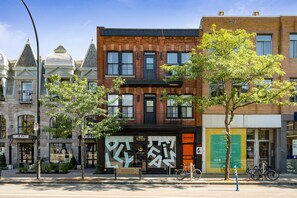 A street view with a row of buildings, including a brick building with a black awning and a storefront with a large geometric sign. - François-Denis Apartments (Montreal)