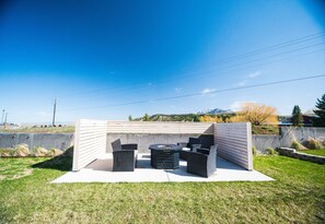 An outdoor patio area with a round table and chairs, surrounded by a low wall and a grassy lawn. - NEW- Modern Home Close to Downtown - Hot Tub! (Bozeman)
