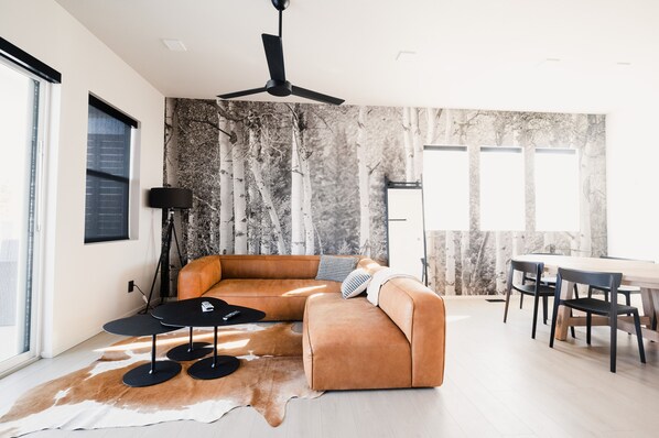 A modern living room with a sectional sofa, a dining table with chairs, a ceiling fan, and a large window. - NEW- Modern Home Close to Downtown - Hot Tub! (Bozeman)