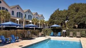 A pool area with lounge chairs and umbrellas, surrounded by a fence and a building with multiple windows. - 2nd row Ocean Views 4br 3ba Sleeps 12, '26 Pool Amenity Card, linens included (Edisto Island)