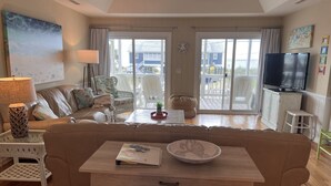 A cozy living room with a sofa, a coffee table, and a TV. There is a balcony with a view of houses. - 2nd row Ocean Views 4br 3ba Sleeps 12, '26 Pool Amenity Card, linens included (Edisto Island)