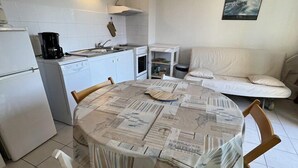 Ett kompakt kök med vita vitvaror, ett matbord täckt med tidningspapper och en soffa i vardagsrummet.