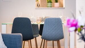 A modern dining area with a table set for four, featuring grey upholstered chairs with wooden legs. Above the table, there are three pendant lights with a geometric design. The wall behind the table displays framed floral artwork. - BoRa Apartment First - Villach (Villach)