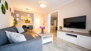 A modern living room with a grey sofa, a coffee table, a flat-screen TV, and a dining area with chairs and a table. - BoRa Apartment First - Villach (Villach)