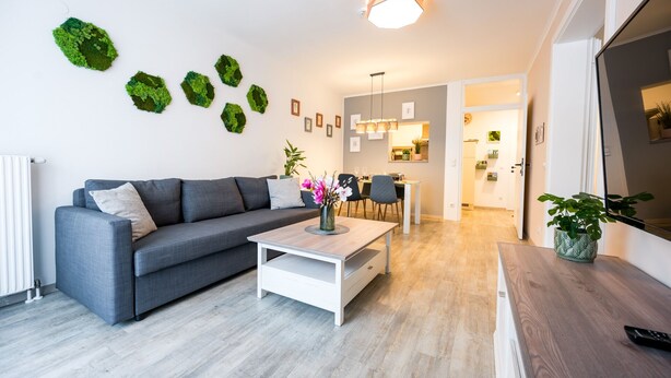 A modern living room with a grey sofa, a white coffee table, and a dining area with a wooden table and chairs. - BoRa Apartment First - Villach (Villach)