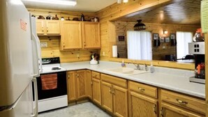 A compact kitchen with wooden cabinets, a white refrigerator, a stove, and a large window with curtains. - Hide a way in our Lodge that sleeps 18! (Indian River)