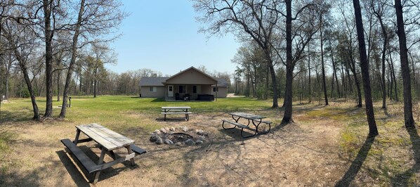 Una cabaña con una fogata y mesas de picnic en un área boscosa.