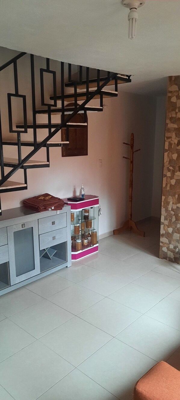 A room with a staircase, a wooden coat rack, a pink and white cabinet with bottles, and a stone wall.