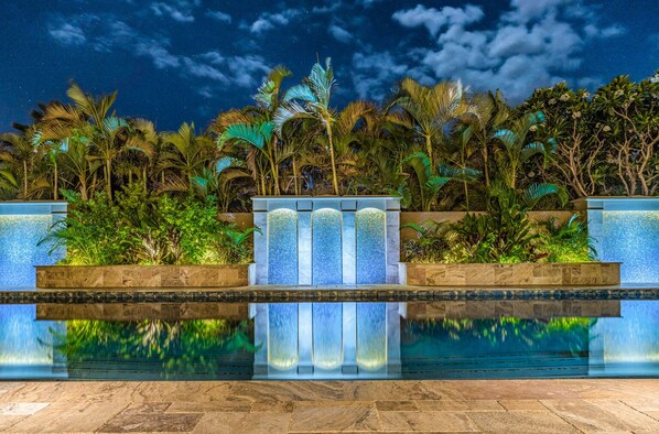 Un espace piscine nocturne où les lumières se reflètent clairement, entouré d’une végétation tropicale luxuriante et agrémenté d’un mur décoratif aux éléments verticaux.
