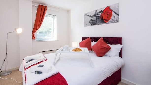 Ein Hotelzimmer mit Bett, Nachttisch mit Essbrett, roher Tagesdecke, Kissen in Rot und Weiß, einem Bild an der Wand und einer Stehlampe.