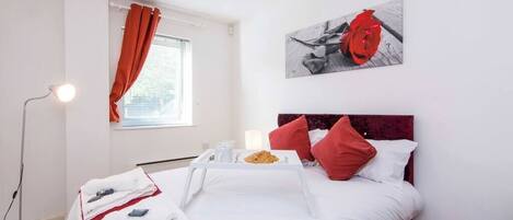 Habitación de hotel con cama, una mesita de noche con bandeja de comida, una manta roja, un juego de almohadas rojo y blanco, un cuadro en la pared y una lámpara de pie.