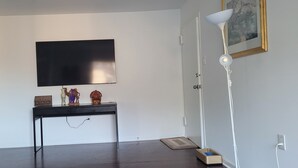 Une pièce dotée d’un téléviseur à écran plat fixé au mur, d’une petite console ornée d’objets décoratifs, d’un lampadaire blanc et d’une porte surmontée d’un tableau encadré.