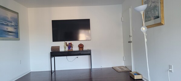 A room with a flat-screen TV mounted on the wall, a small console table with decorative items, a white floor lamp, and a door with a framed picture above it.