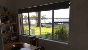 A dining area with a table and chairs, a bookshelf, and a window with a view of a body of water and a bridge. - “Mona’s On Rockpool” - Waterfront heaven (Tuncurry)