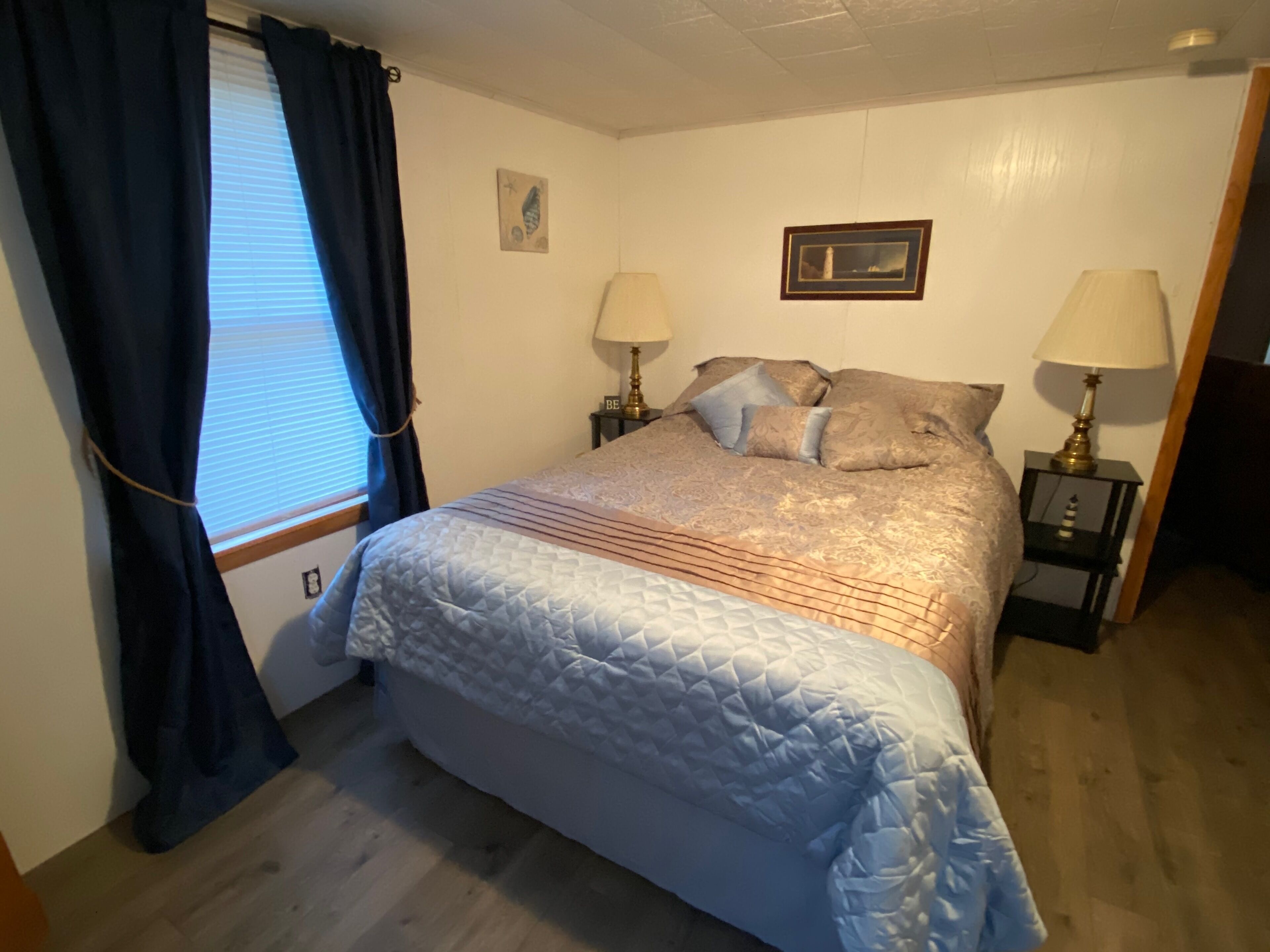 Un dormitorio con cama, dos mesitas de noche con lámparas, una ventana con cortinas y un cuadro colgado en la pared.
