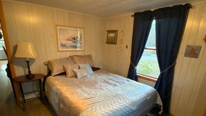 A bedroom with a bed, a nightstand, a lamp, a painting on the wall, and a window with curtains. - Cottage near the Bay (Crisfield)