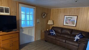 A living room with a brown leather couch, wooden walls, a television on a wooden cabinet, a door with blinds, and a framed picture on the wall. - Cottage near the Bay (Crisfield)