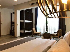 Une chambre d’hôtel avec un grand lit, un lit superposé, deux chaises, une petite table et un luminaire suspendu.