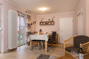 Un coin repas avec une table dressée pour deux personnes, une chaise en osier et une bouteille de vin posée sur une petite table d’appoint.