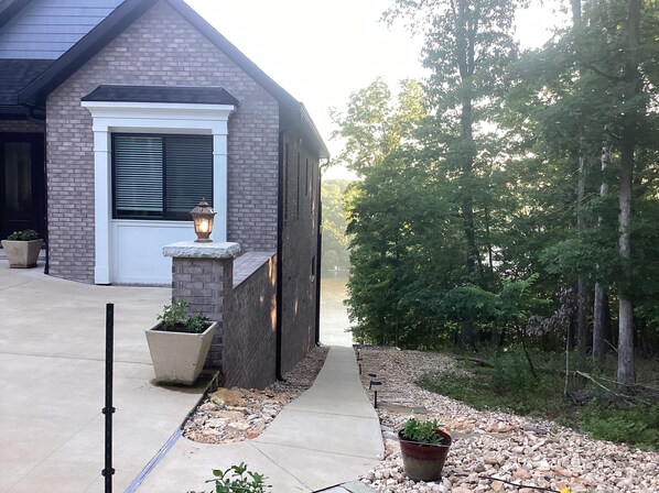 A house with a brick exterior, a white door, and a stone ledge with a lantern. There are potted plants and a pathway leading to a body of water.