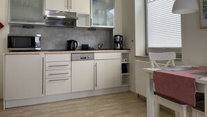 A compact kitchen with white cabinets, a microwave, a coffee maker, and a window with blinds. - Strandhausvilla Wo. 2 - "Strandhausvilla - Whg.2" Beach chair included! (Dahme)