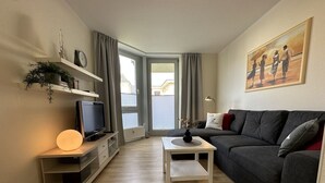 A modern living room with a dark grey sofa, a flat-screen TV on a wooden stand, a white coffee table, and a painting on the wall. - Strandhausvilla Wo. 2 - "Strandhausvilla - Whg.2" Beach chair included! (Dahme)