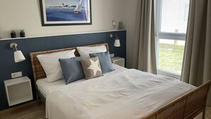 A bedroom with a bed, two bedside tables, a wall-mounted lamp, a framed painting, and a window with curtains. - Strandhausvilla Wo. 2 - "Strandhausvilla - Whg.2" Beach chair included! (Dahme)
