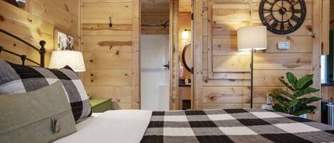 Una habitación de madera con una cama, una mesita de noche, una lámpara, un reloj de pared y una puerta que da a otra habitación.