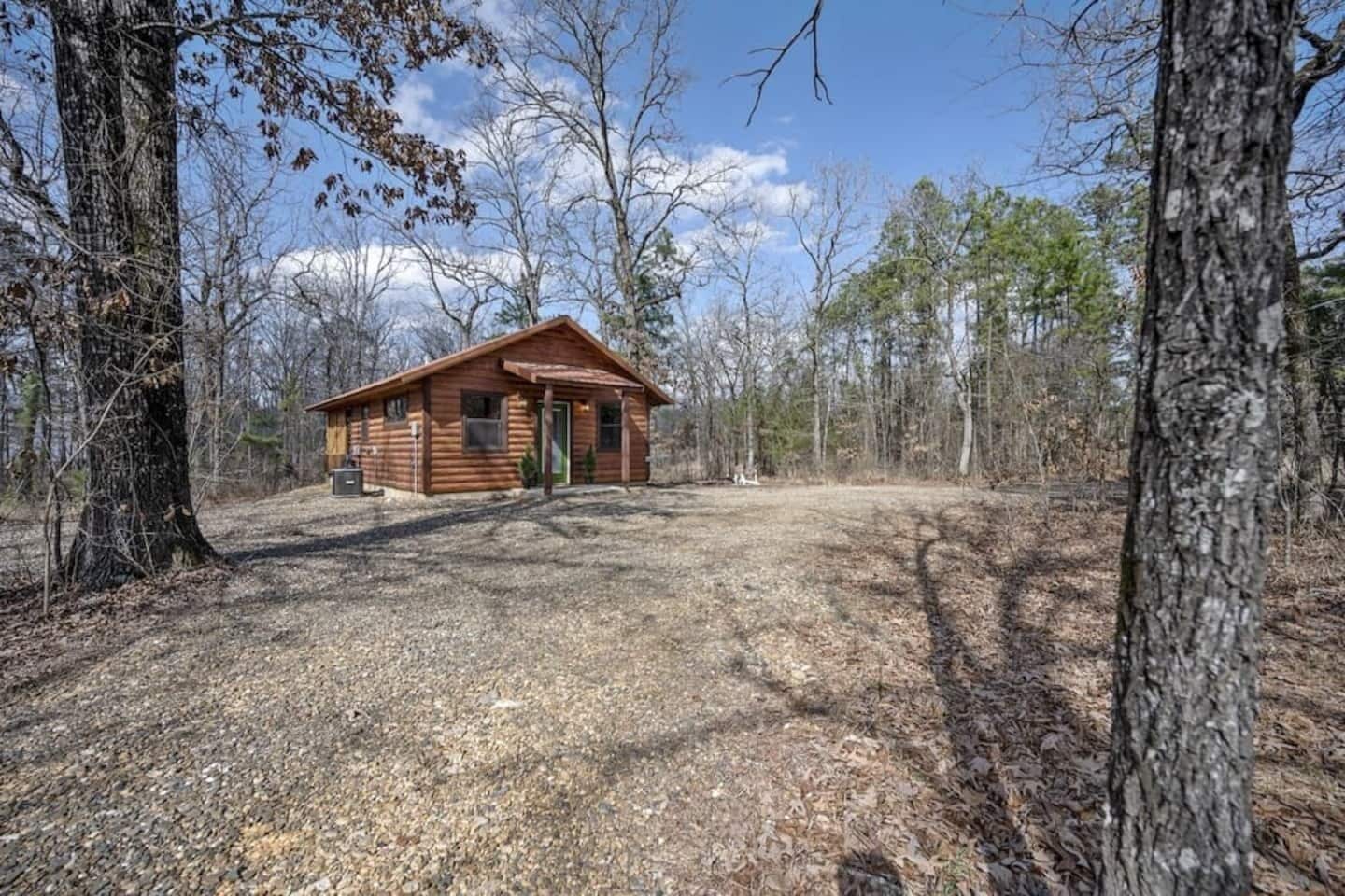 Une petite cabane en rondins avec une porte verte, entourée d’arbres et bordée d’une allée de gravier.