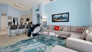 A modern living room with a sofa, coffee table, and a massage chair. There is a kitchen area with a bar and chairs.