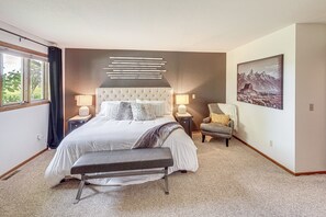 A bedroom with a large bed, bedside tables, a bench, a chair, a painting, and a window with curtains.