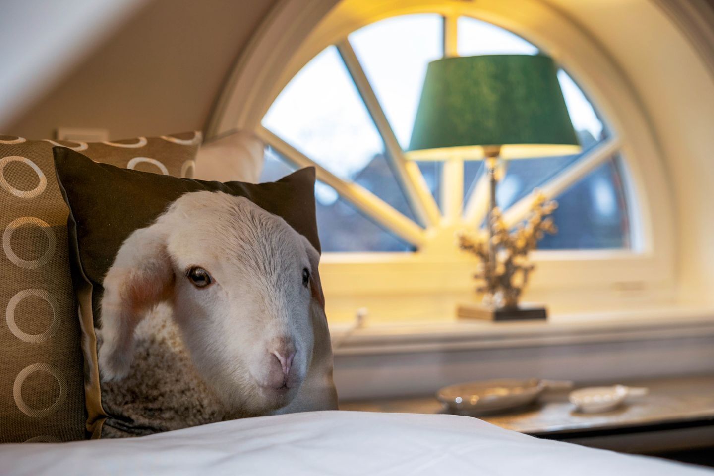 Un lit avec un oreiller orné d’un motif représentant la tête d’un mouton, une lampe et une fenêtre donnant sur le ciel.
