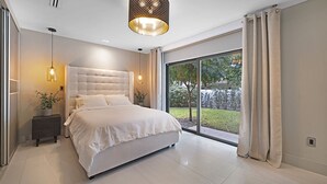 A modern bedroom with a large bed, a bedside table, a potted plant, and a view of a garden through a sliding door. - Private Saltwater Pool - Updated Modern 3BR 3BA - 1 Mile to Beach! (Fort Lauderdale)