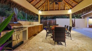 Un área de cocina al aire libre con techo de paja, piso de piedra y un juego de comedor.