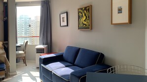 A modern living room with a blue sofa, a dining table, and a chair. There are two framed artworks on the wall and a window with curtains. - Akile B. Ribeiro (São Paulo)
